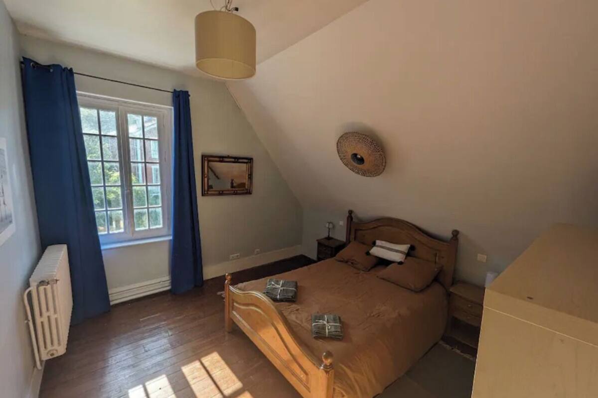 Un dormitorio con cama de madera, una ventana con cortinas, un radiador, una lámpara de pared y un cuadro enmarcado.