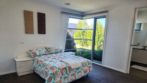 A bedroom with a large bed, a white nightstand, a window with a view of trees, and a bathroom with a sink and mirror. - Tranquil in Alfredton (Alfredton)