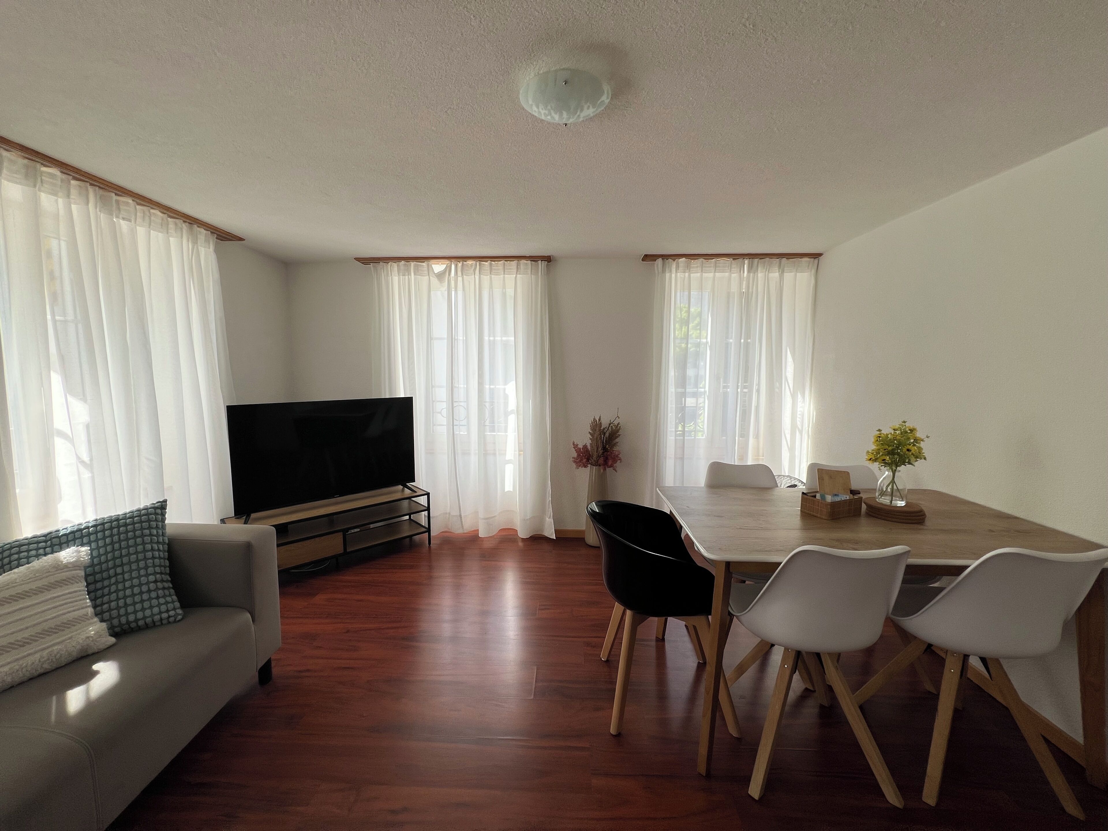 Sala de estar com sofá, televisão, mesa de jantar com cadeiras e cortinas leves.