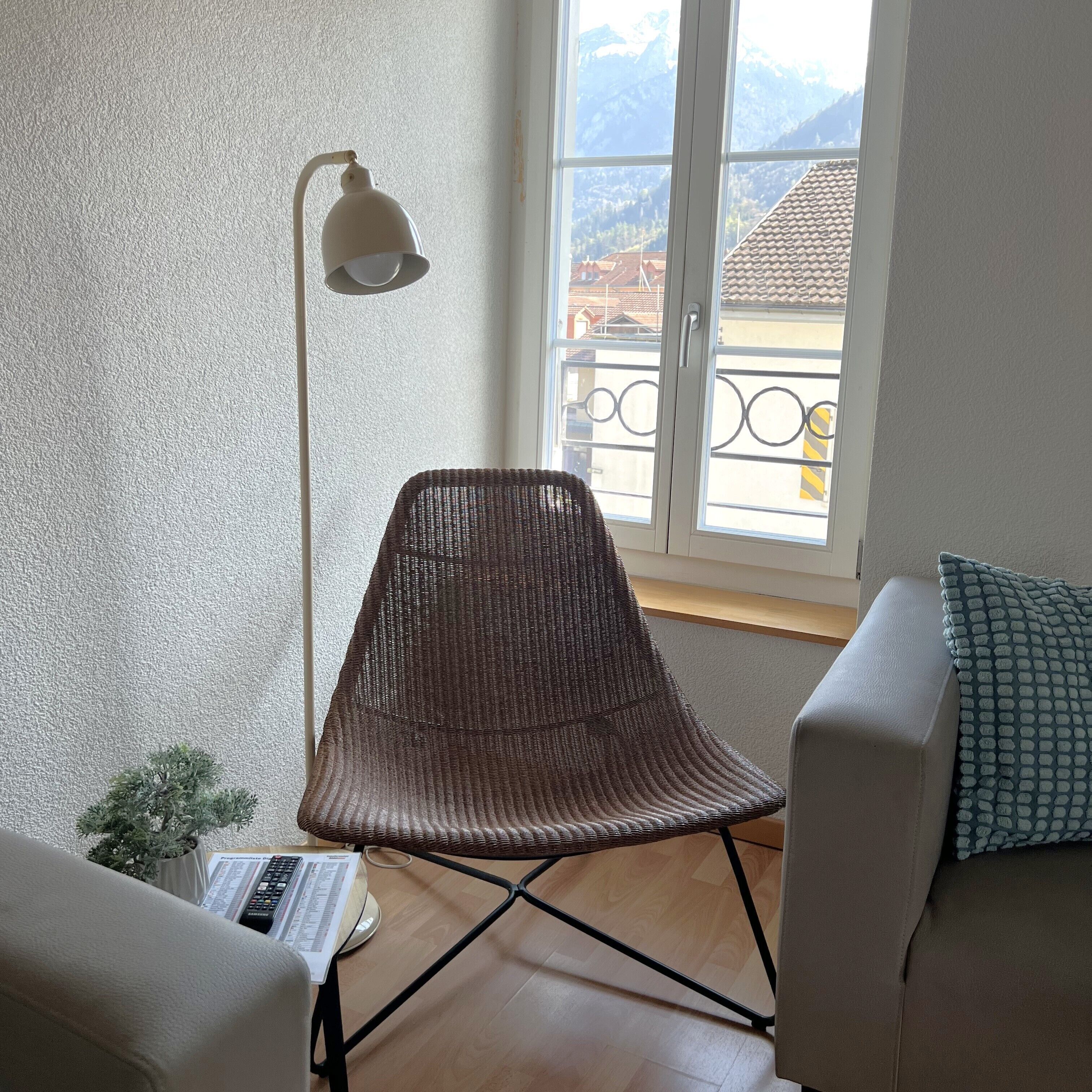 A room with a wicker chair, a white sofa, a standing lamp, and a view of mountains through a window.