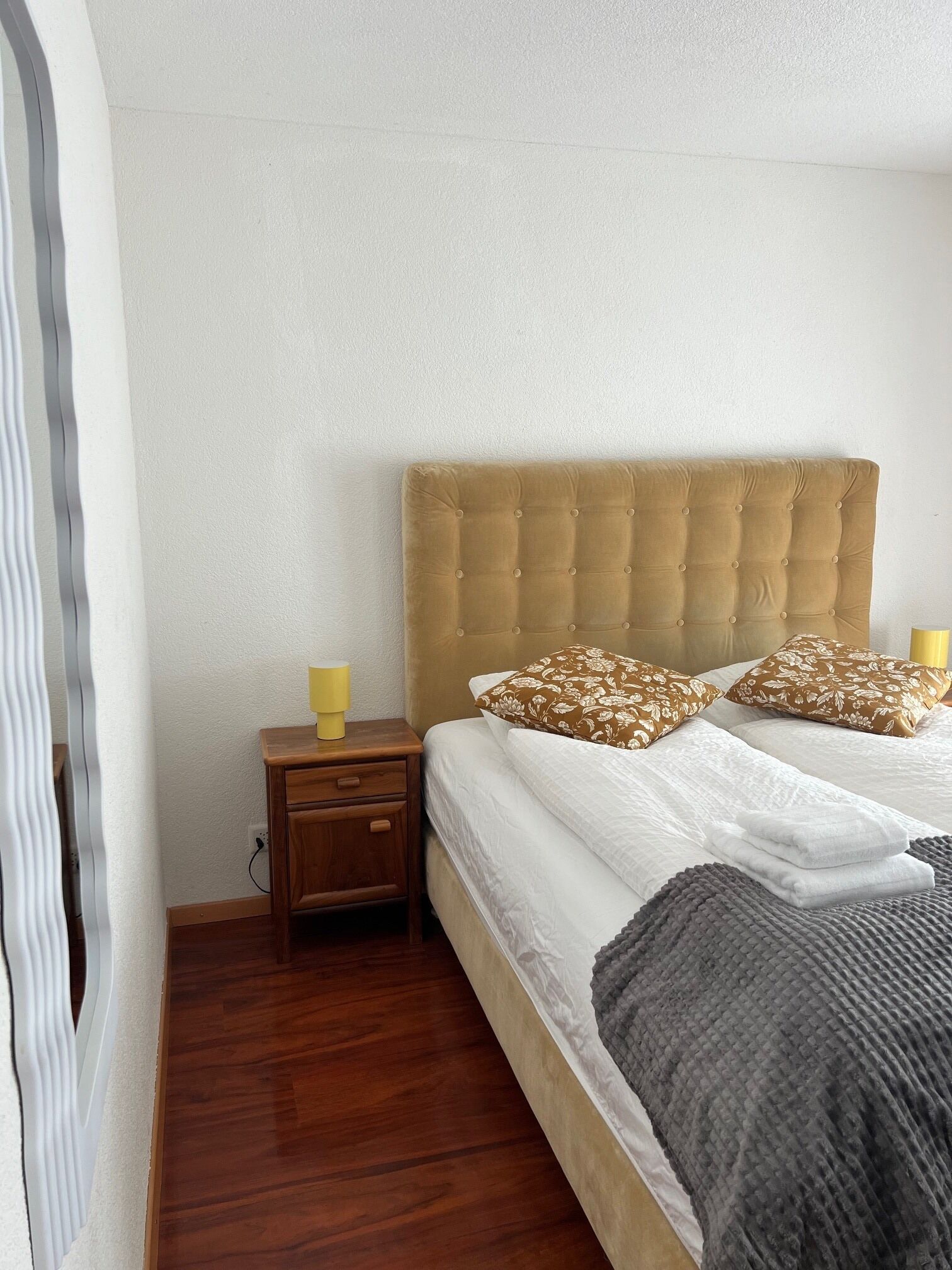 A bedroom with a tufted headboard, a wooden nightstand with a lamp, two beds with floral pillows, and a grey blanket.