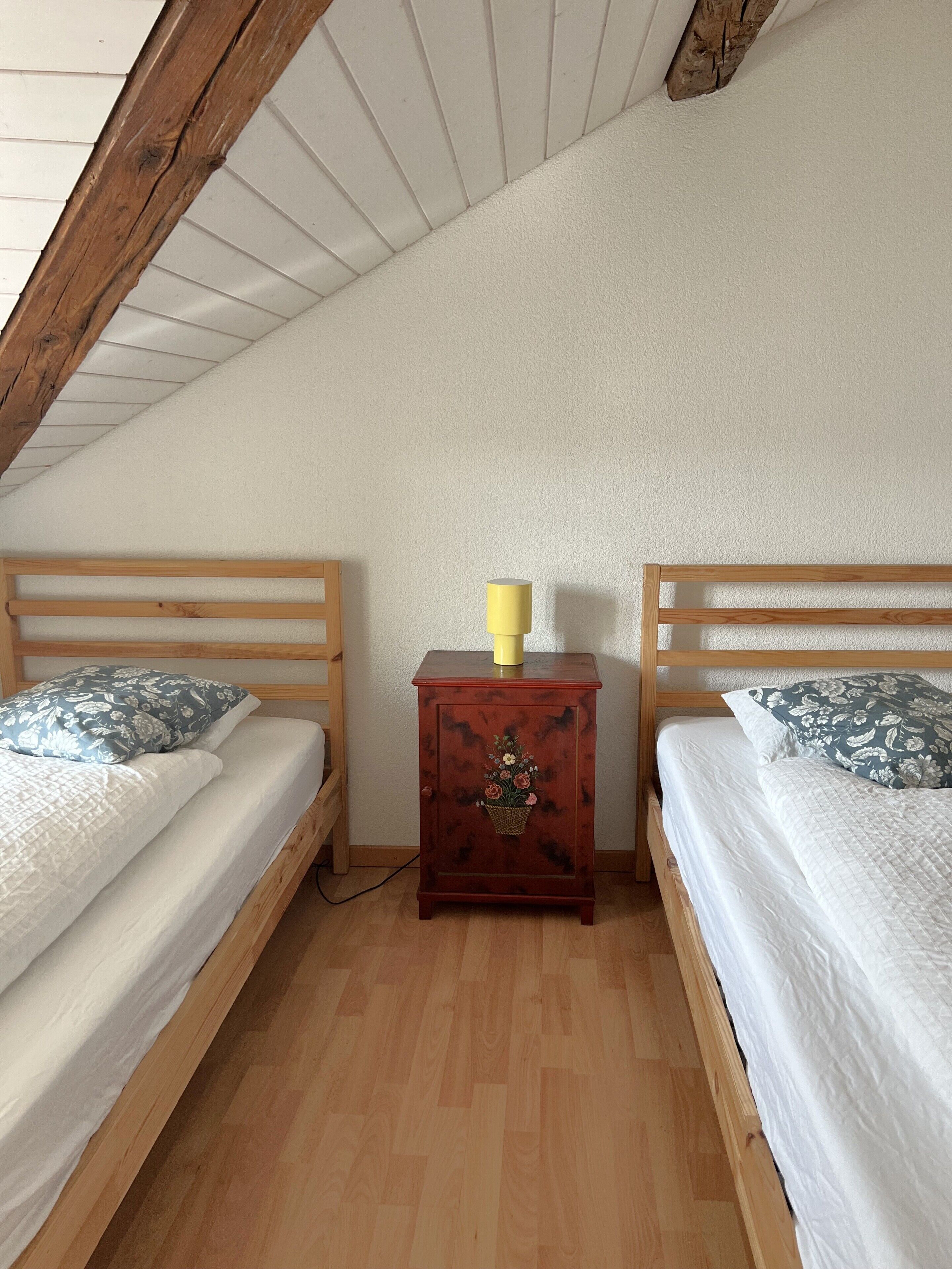 A bedroom with two wooden beds, a small wooden nightstand with a yellow lamp, and a wooden ceiling.