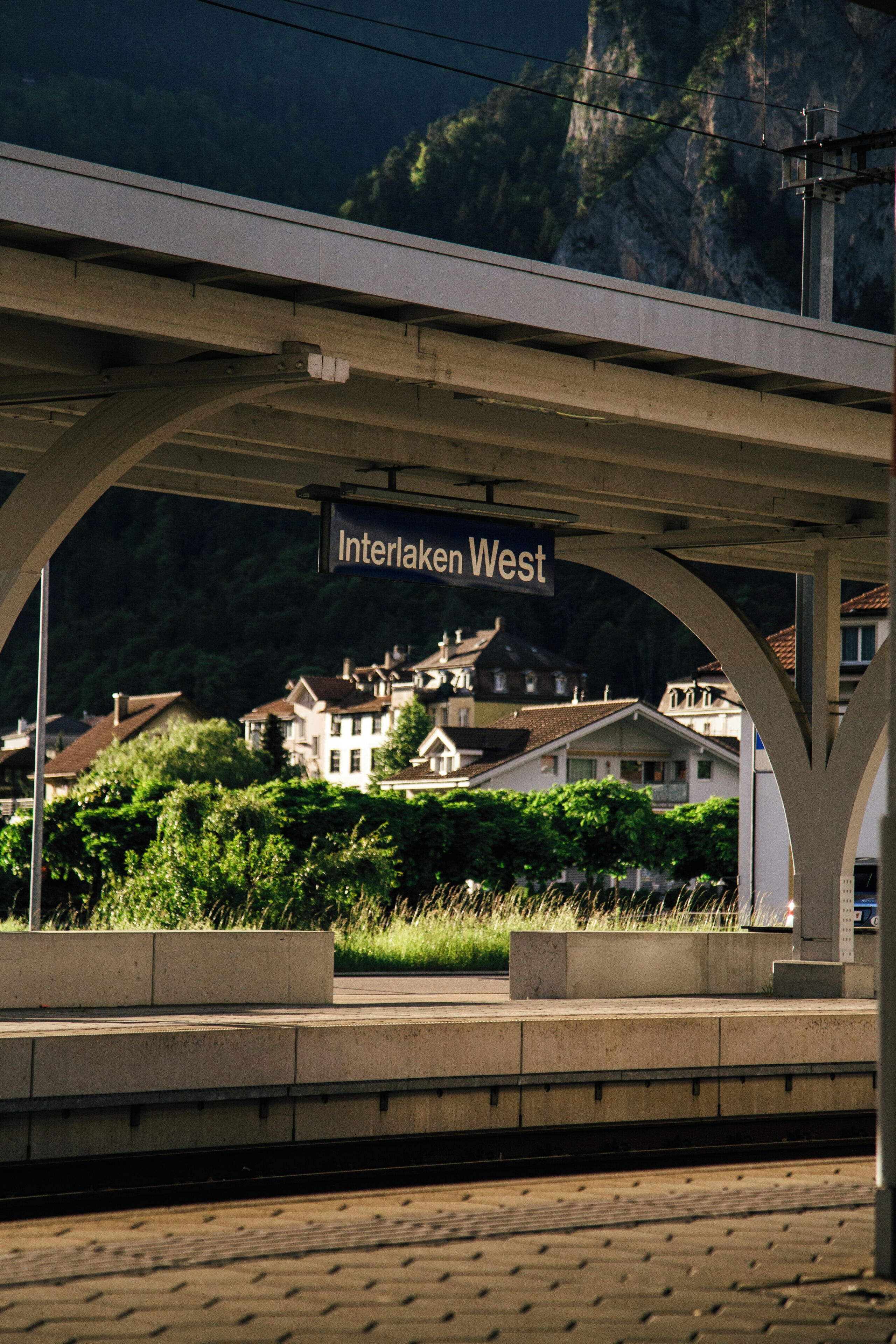 A train station platform with a sign reading 'Interlaken West' and a view of houses in the background.