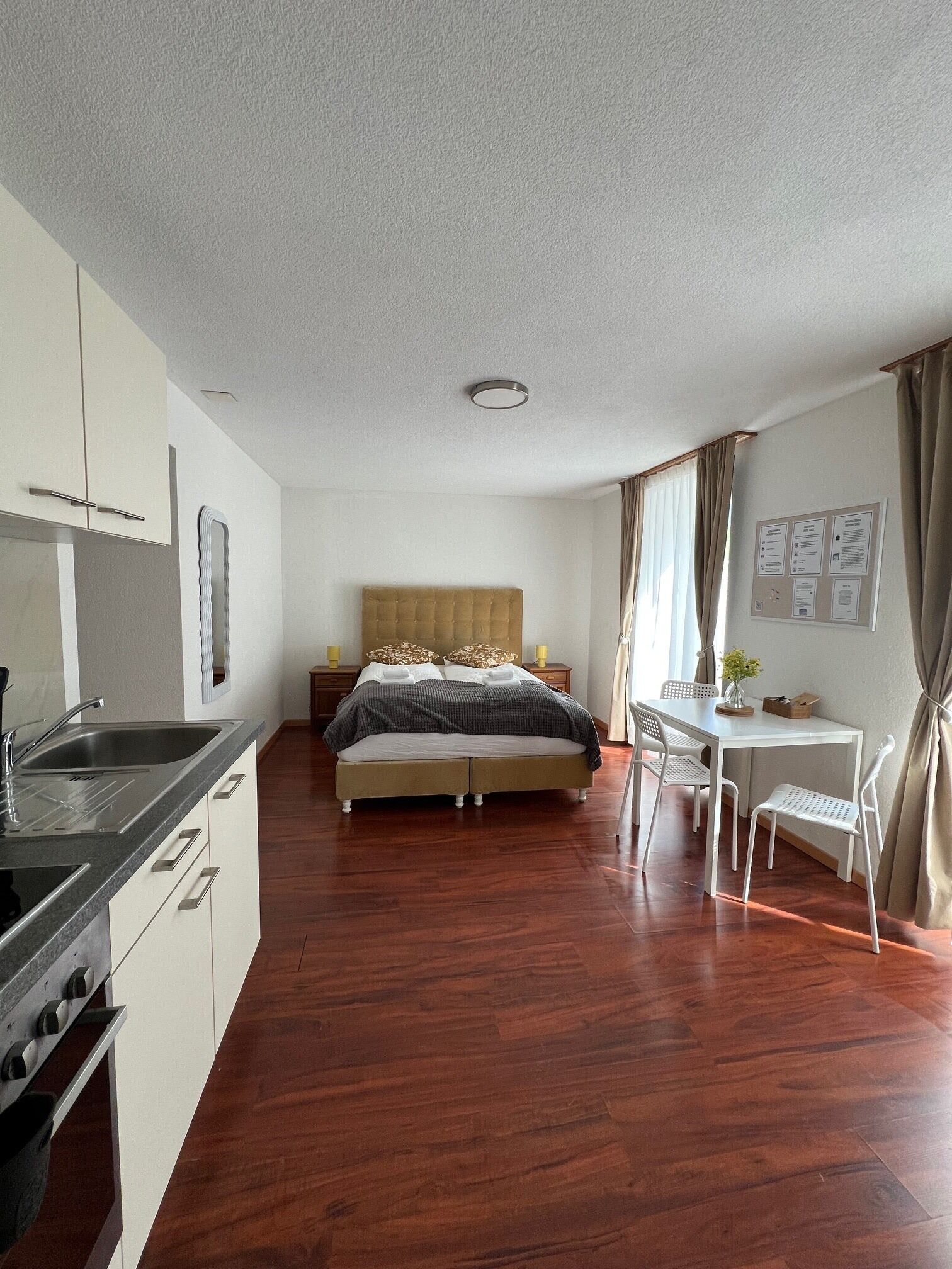 A modern hotel room with a wooden floor, a bed with a headboard, a desk with chairs, a small table with a vase, and a wall-mounted bulletin board.