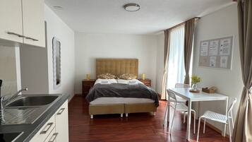 A modern hotel room with a wooden floor, a bed with a headboard, a desk with chairs, a small table with a vase, and a wall-mounted bulletin board.