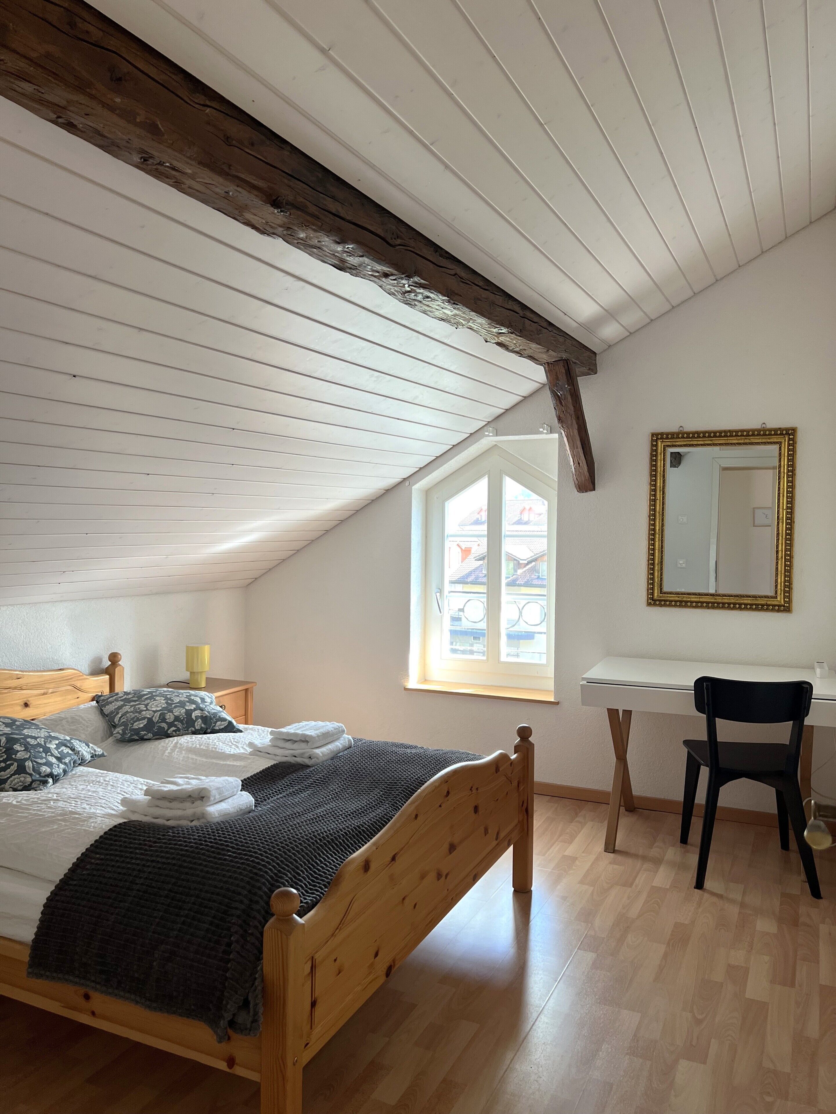 Ein Zimmer mit einem hölzernen Bett, einem Schreibtisch, einem Stuhl, einem Fenster und einem Spiegel.