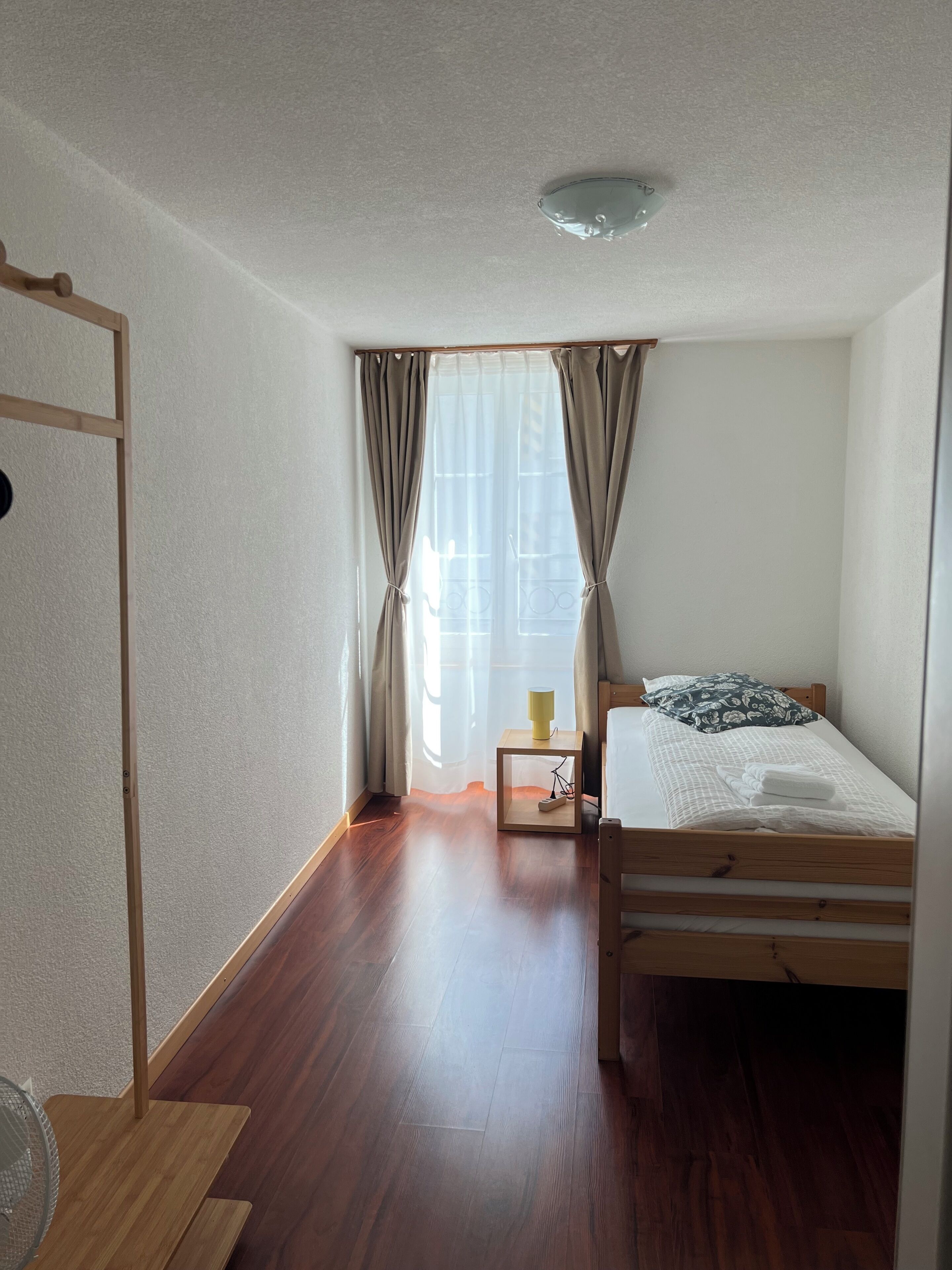 Een kamer met een houten bed, een klein nachtkastje met een lamp, een ventilator en een raam met gordijnen.