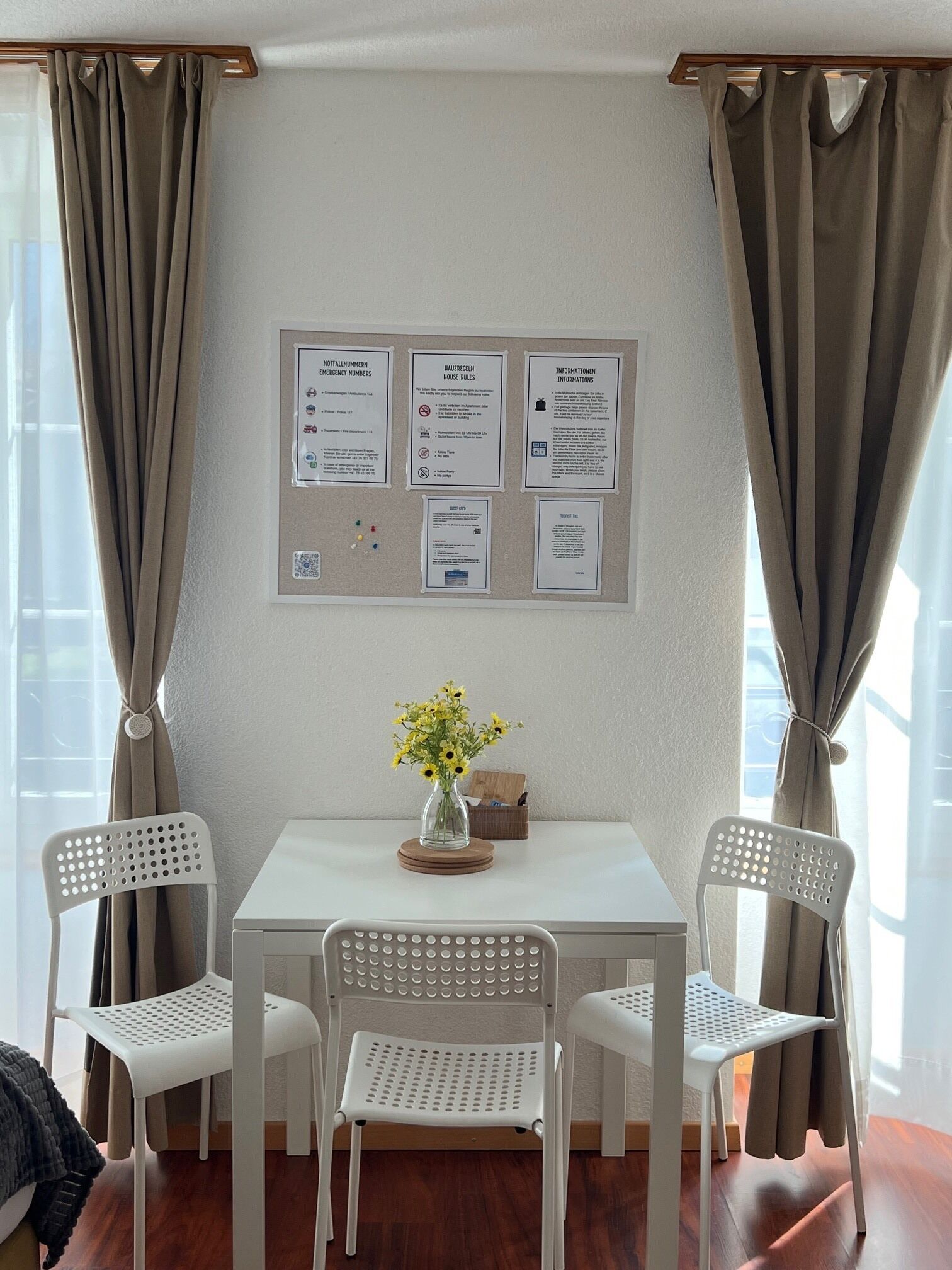 A room with a table, four chairs, a vase with flowers, and a bulletin board with emergency numbers and house rules.