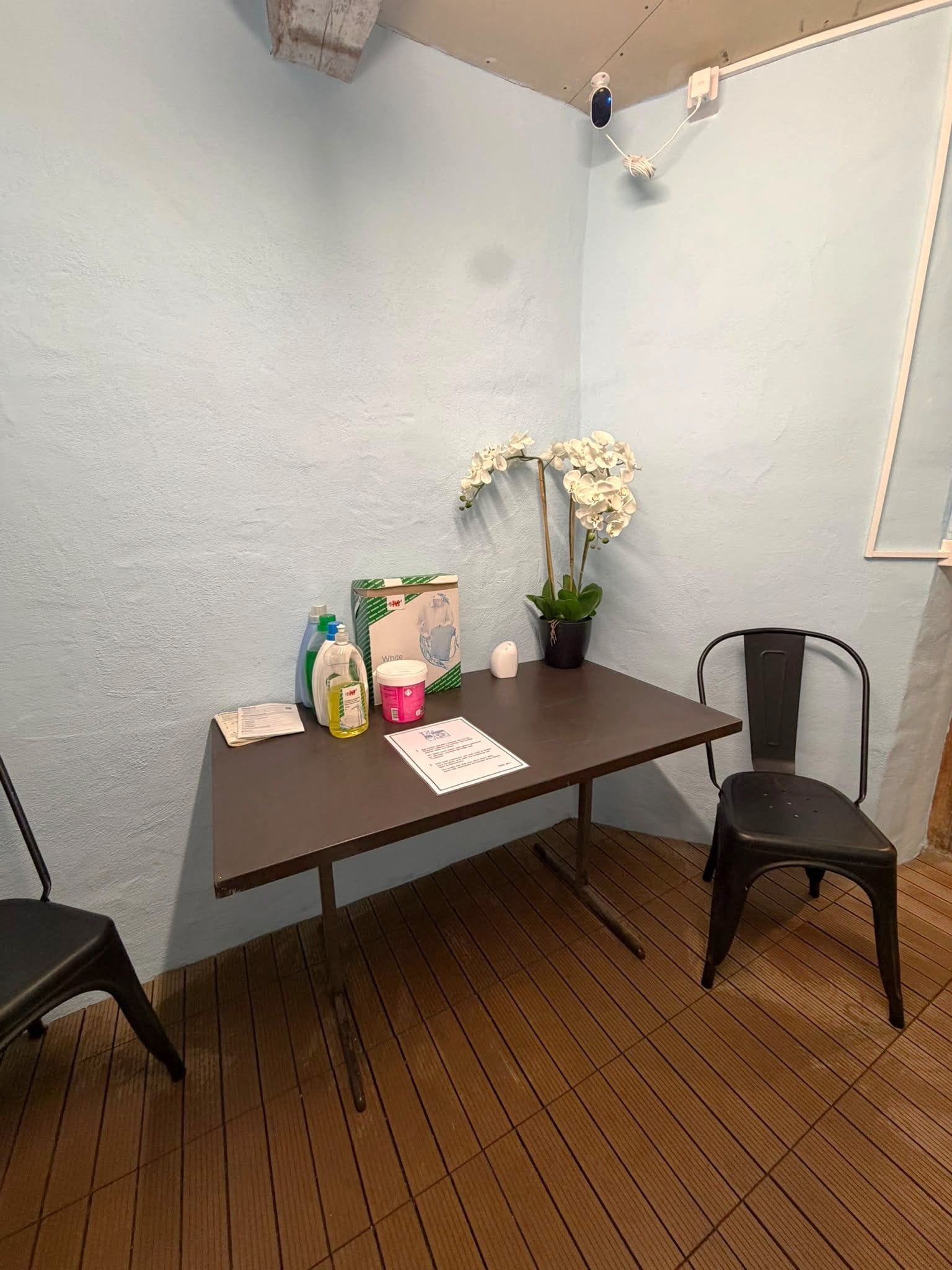Una habitación pequeña con piso de madera, una mesa con una silla, una planta en maceta y algunos productos de limpieza.