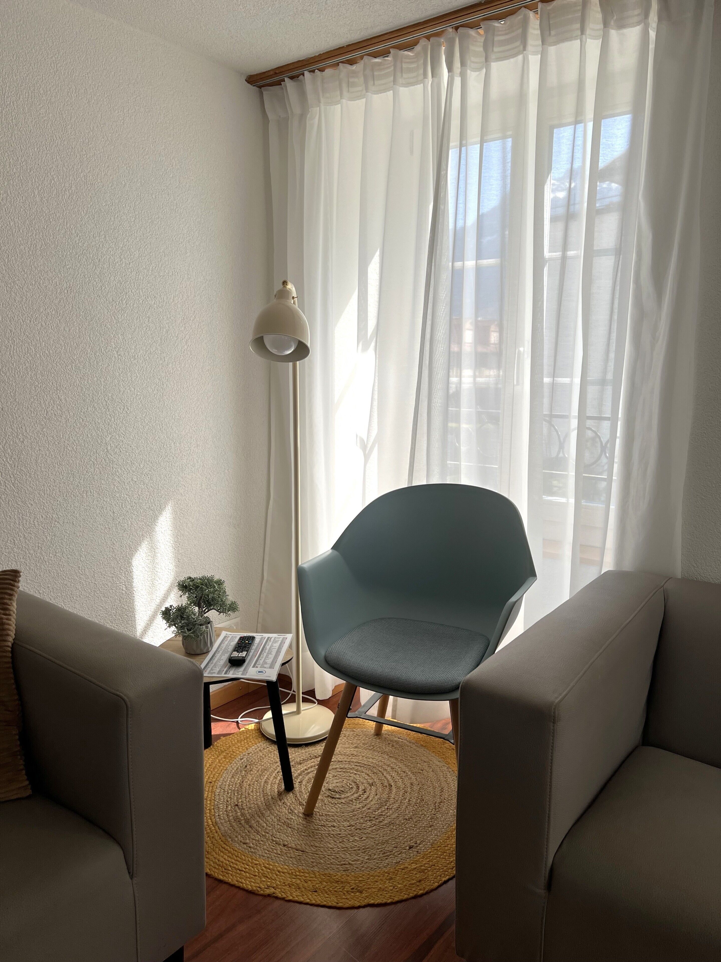 A room with a modern chair, a small table with a plant, a lamp, and a view through sheer curtains.