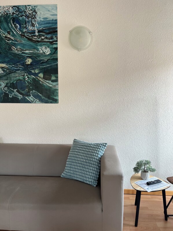 Una sala de estar con un sofá gris, un cojín estampado, una lámpara fijada a la pared y una mesita con un bonsái y un control remoto.