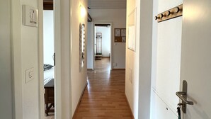 A hallway with wooden flooring, white walls, and a ceiling with exposed beams. There are light switches and a door with a handle on the right side. - Nimbuzzz Apartments (Unterseen)
