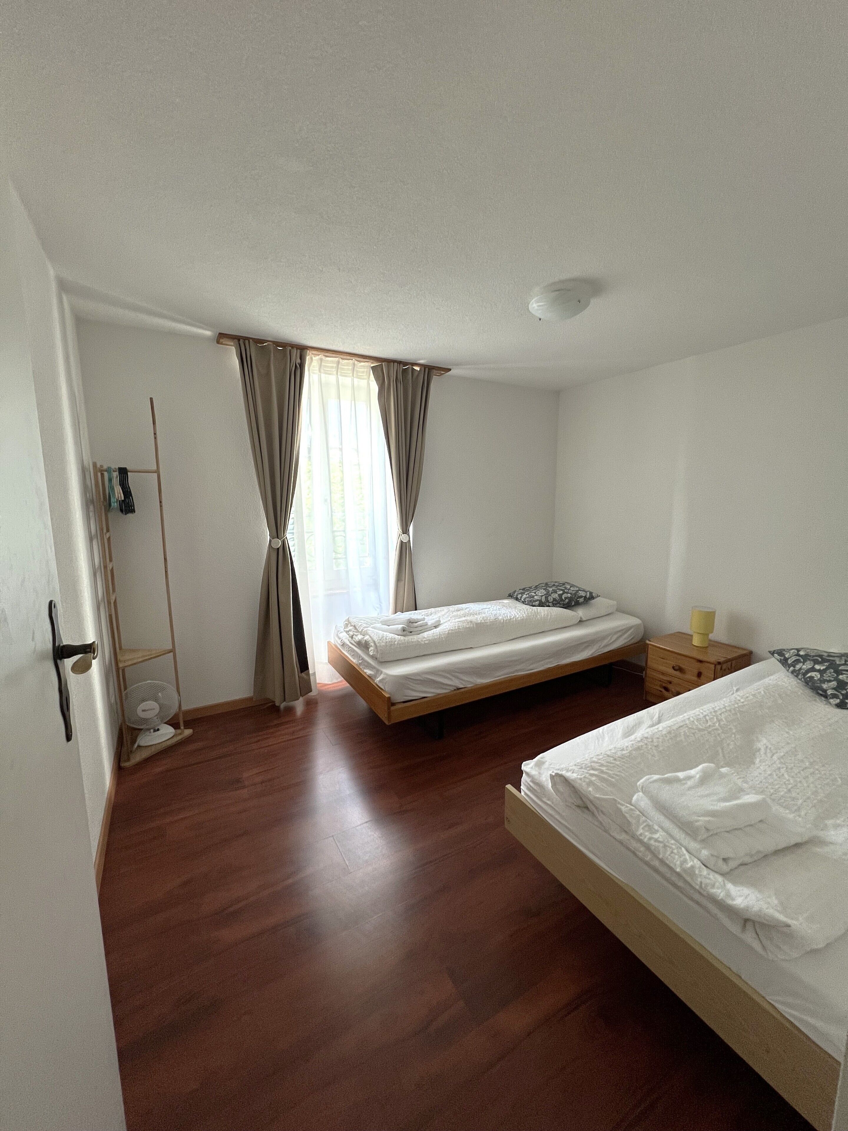 Una habitación con dos camas, piso de madera, un ventilador y una ventana con cortinas.