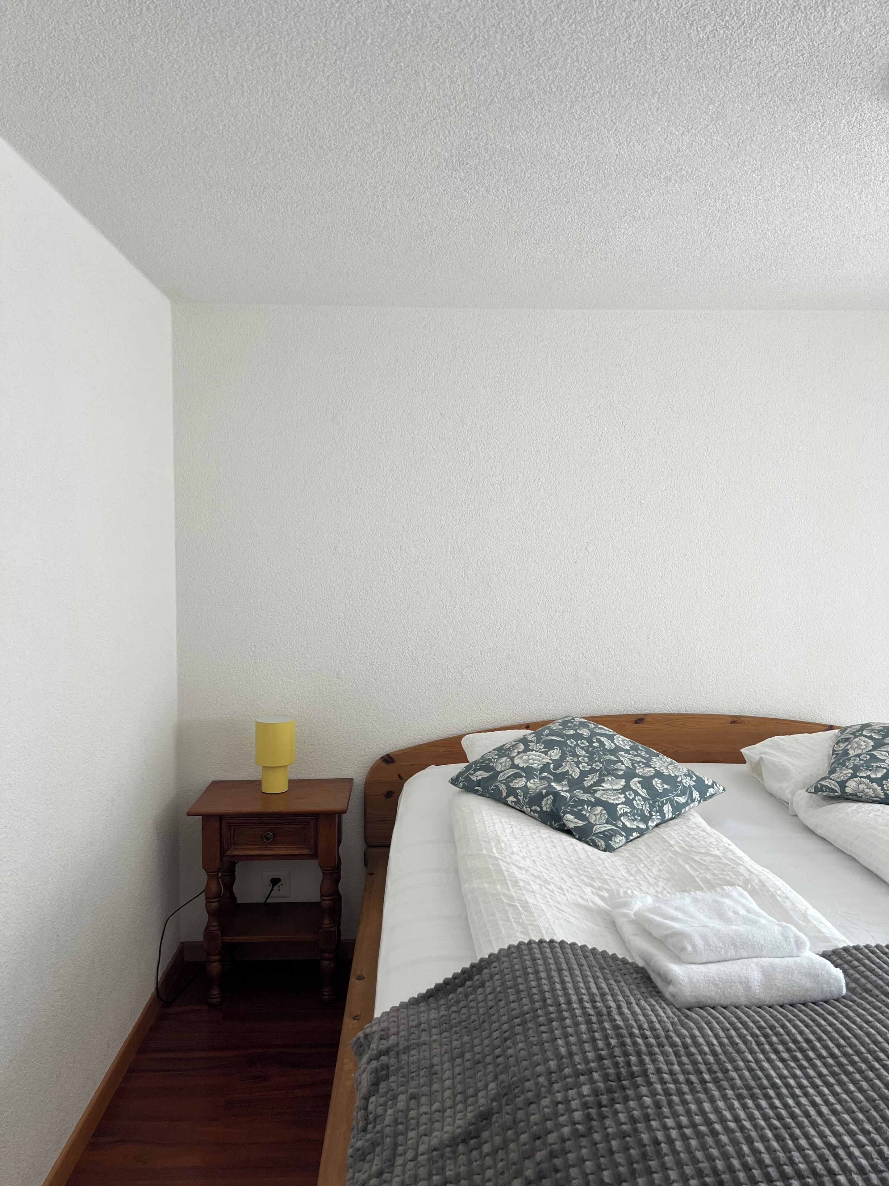 A neatly made bed with a grey blanket and floral pillows, a wooden nightstand with a lamp, and a wooden floor.