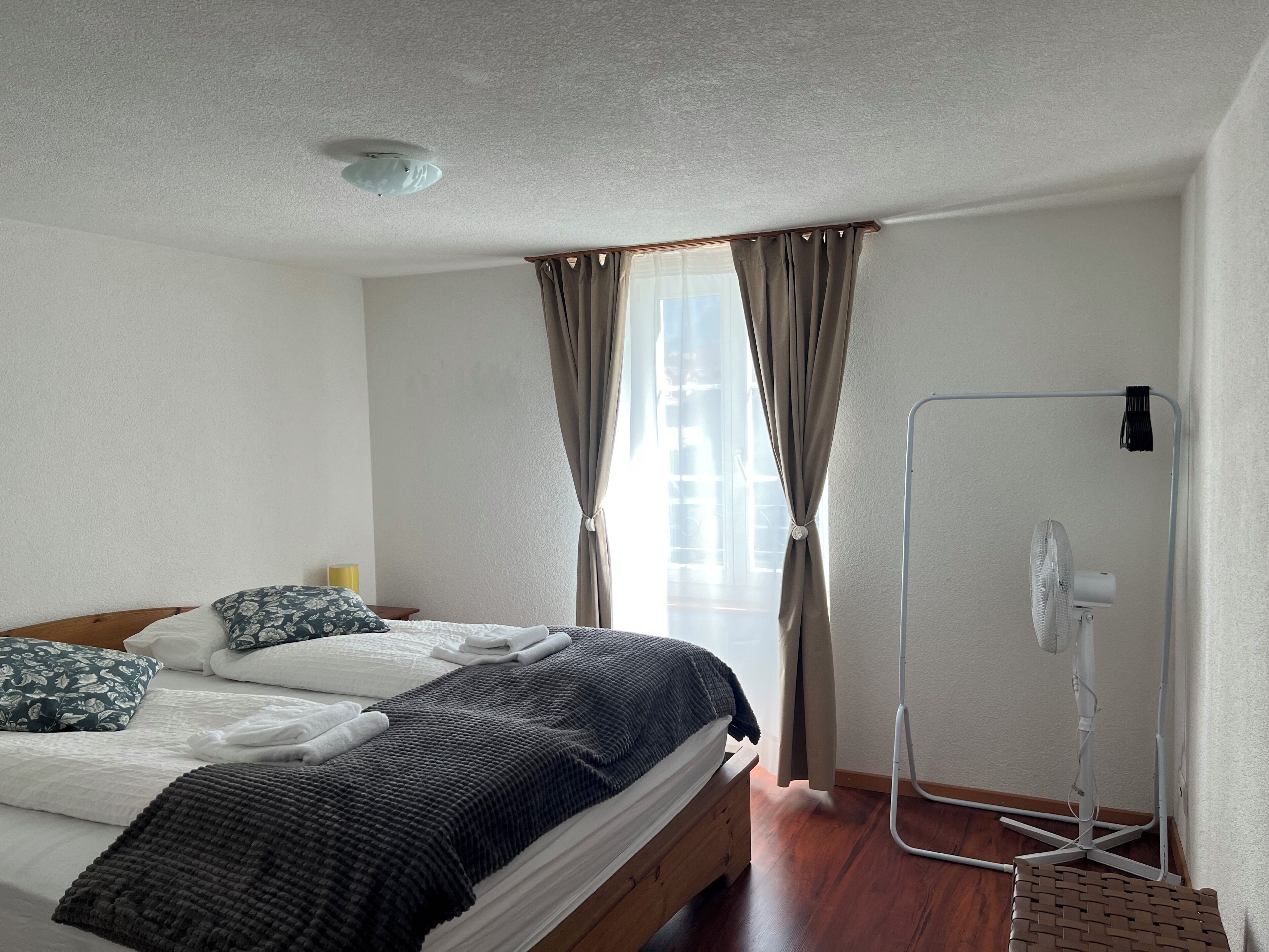 Una cama bien hecha con almohadas, un ventilador de pie y una ventana con cortinas.
