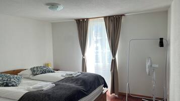 Una cama bien hecha con almohadas, un ventilador de pie y una ventana con cortinas.