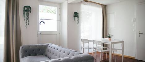 Ein Zimmer mit grauem Sofa, einem weißen Esstisch mit Stühlen und hängenden Pflanzen.