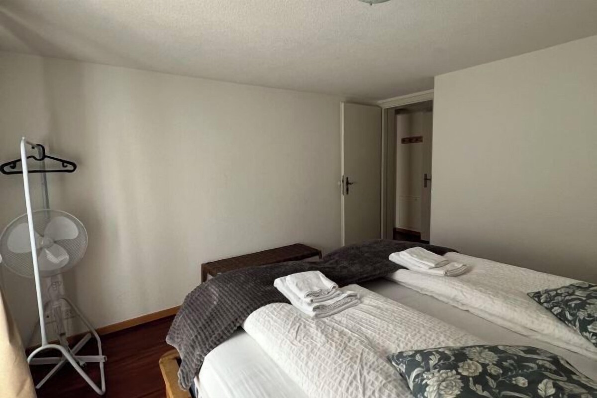 Une chambre avec deux lits, un ventilateur sur pied et un porte-manteau.