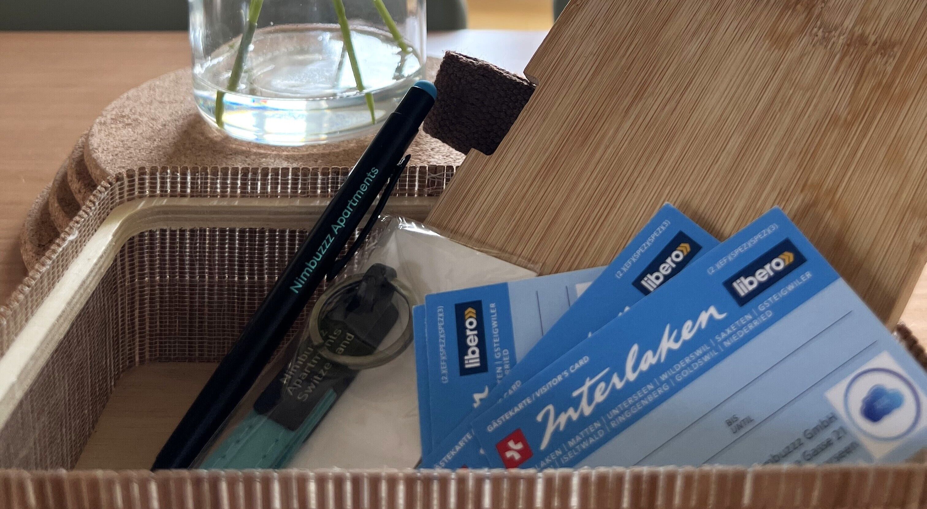 Una cesta de mimbre con un bolígrafo negro, una tarjeta azul y un pequeño recipiente de vidrio.