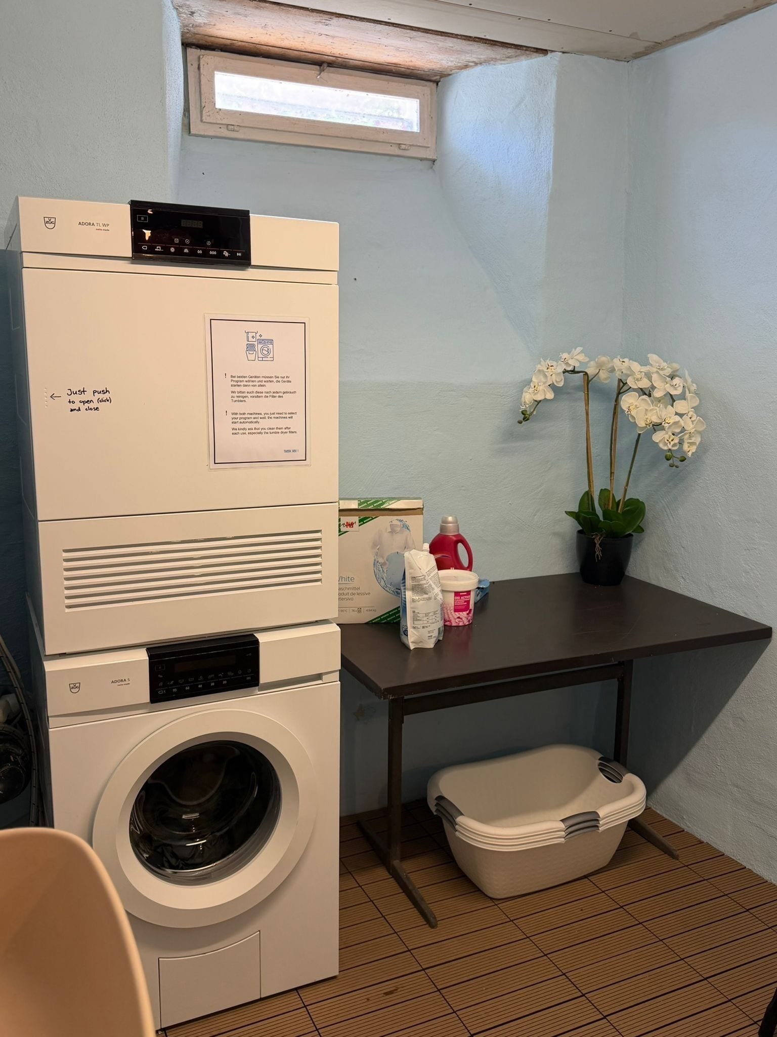 A laundry room with a stacked washer and dryer, a small table with cleaning supplies, a potted orchid, and a window.