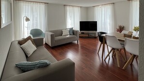 A modern living room with a sofa, TV, dining table, and chairs. - Nimbuzzz Apartments (Unterseen)