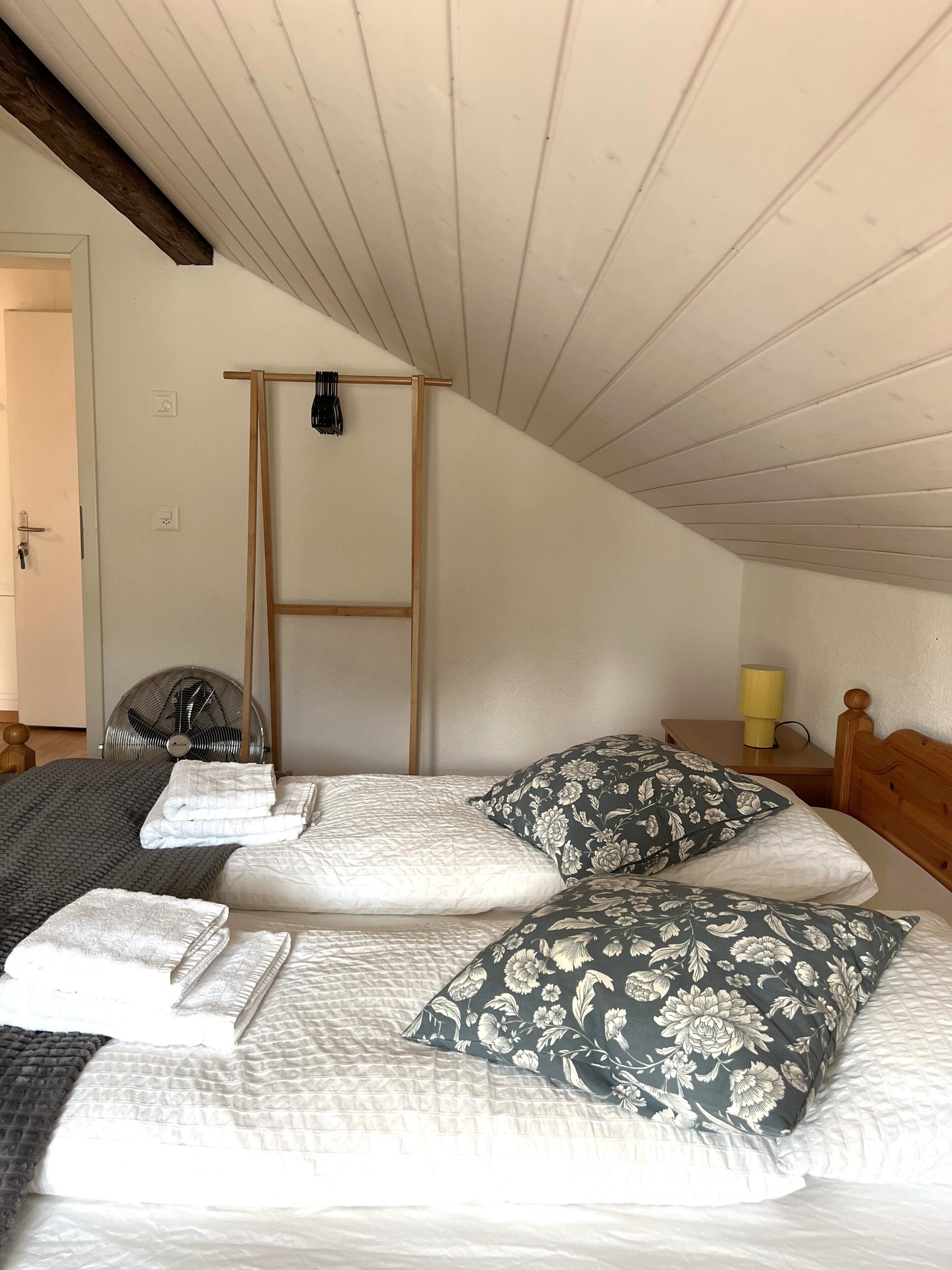 Een slaapkamer met een hoogslaper, een houten trapje, een ventilator en een nachtkastje met lamp.