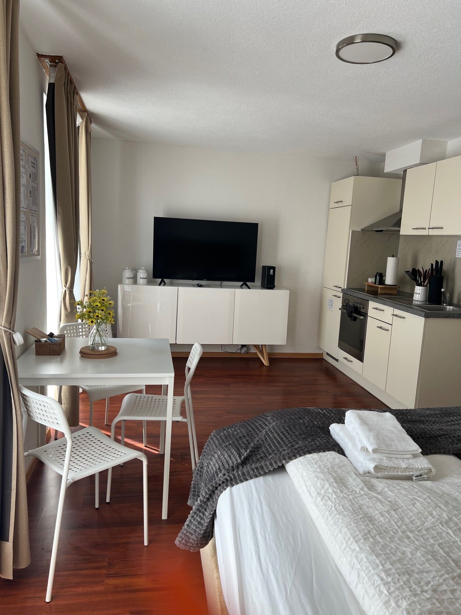 Camera d'albergo con un letto, una TV, un tavolo da pranzo con sedie, un angolo cottura e una finestra con tende.
