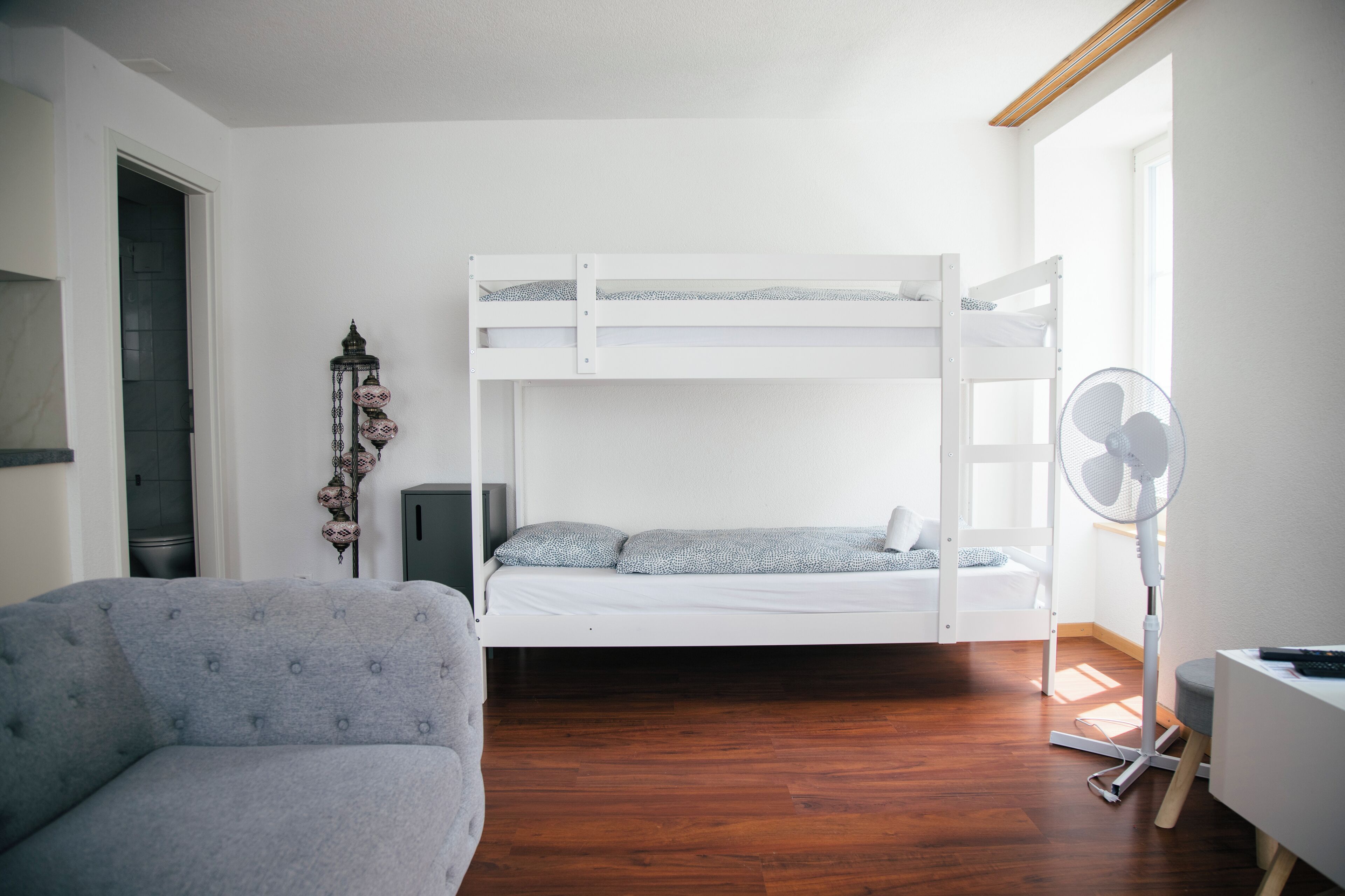 A room with a bunk bed, a grey sofa, a standing fan, and a decorative lamp.