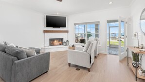 A spacious living room with a fireplace, a flat-screen TV mounted above it, and a large window offering a view of the outdoors.