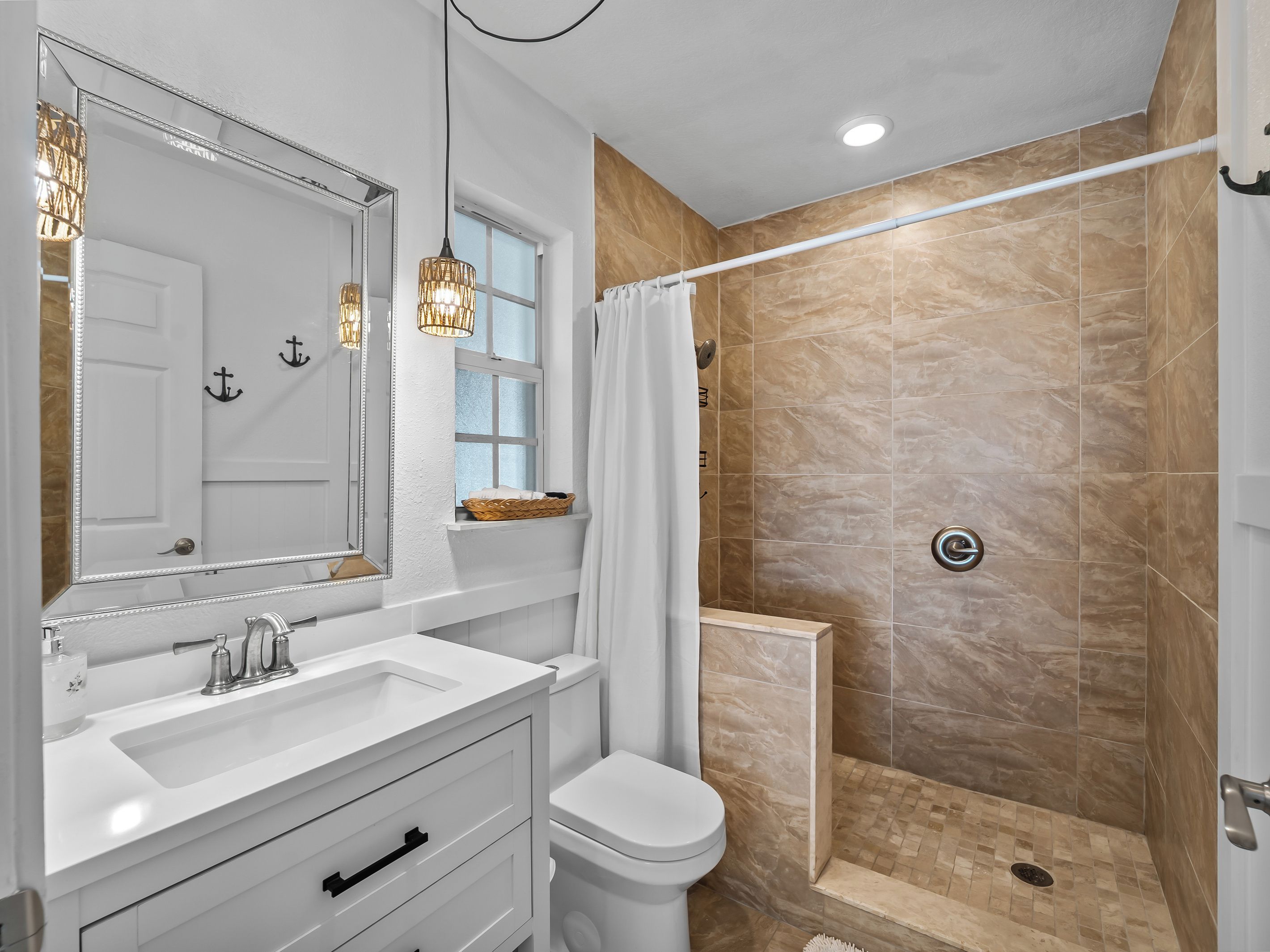 Un baño con tocador blanco, un espejo grande, ducha con azulejos color beige y una ventana con una cesta en el alféizar.