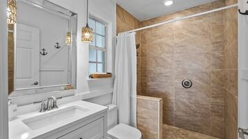 Un baño con tocador blanco, un espejo grande, ducha con azulejos color beige y una ventana con una cesta en el alféizar.