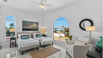 A hotel room with two beds, a ceiling fan, a large window with a view, a round mirror, and a side table with a lamp.