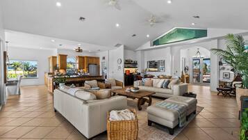 A spacious living area with a large sofa, a coffee table, and a dining table. There is a kitchen in the background with wooden cabinets and a refrigerator.
