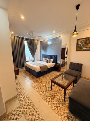 A hotel room with a bed, a sofa, a glass coffee table, patterned floor tiles, a ceiling fan, and a painting on the wall.