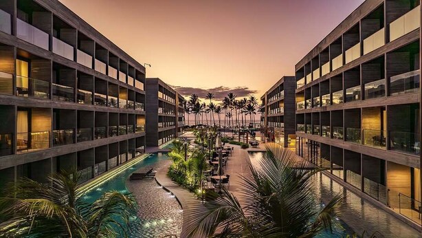 A hotel with a central pool surrounded by palm trees and outdoor seating, featuring multiple floors with balconies. - Porto alto resort  (Ipojuca)