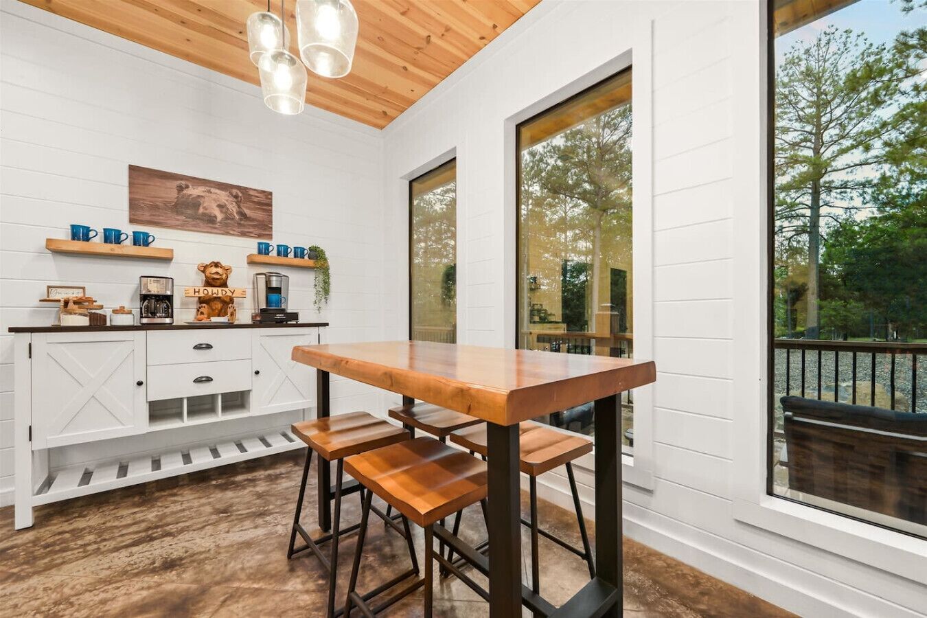 Een keuken met een houten tafel, barstoelen, witte kasten en een groot raam met uitzicht op bomen.