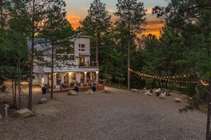 A two-story cabin with a deck and outdoor seating area, surrounded by trees and a gravel ground. - The Grinning Bear|Sleeps 14|Basketball|Pickleball (Broken Bow)
