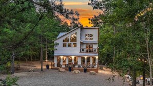 A two-story cabin with a deck, surrounded by trees and a gravel driveway. - The Grinning Bear|Sleeps 14|Basketball|Pickleball (Broken Bow)
