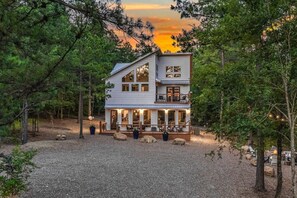 A two-story cabin with a deck, surrounded by trees and a gravel driveway.