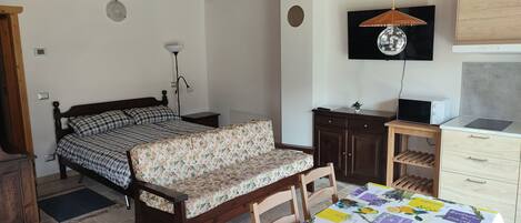 A bedroom with a bed, a desk, a chair, a table, a TV, and a kitchenette.