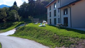 A two-story building with a paved driveway, a grassy lawn with lounge chairs, and a mountain backdrop. - Ai Monti (Barcis)