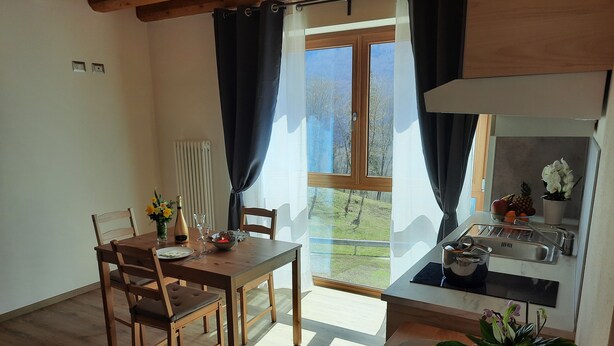 A dining area with a wooden table and chairs, a window with curtains, a kitchen area with a sink and countertop, and a potted plant on the table. - Ai Monti (Barcis)