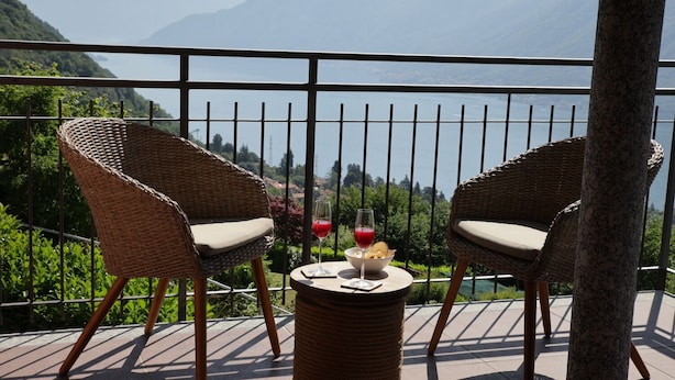 Two wicker chairs with cushions on a balcony with a view of mountains and a lake.