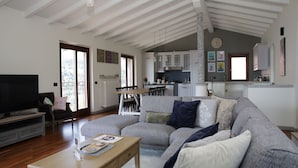 A spacious living room with a large sectional sofa, a wooden coffee table, and a flat-screen TV. There is a dining area with a table and chairs, and a kitchen in the background.