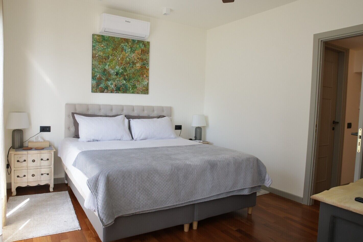 A bedroom with a bed, bedside tables, a nightstand, a wall-mounted air conditioner, and a door.