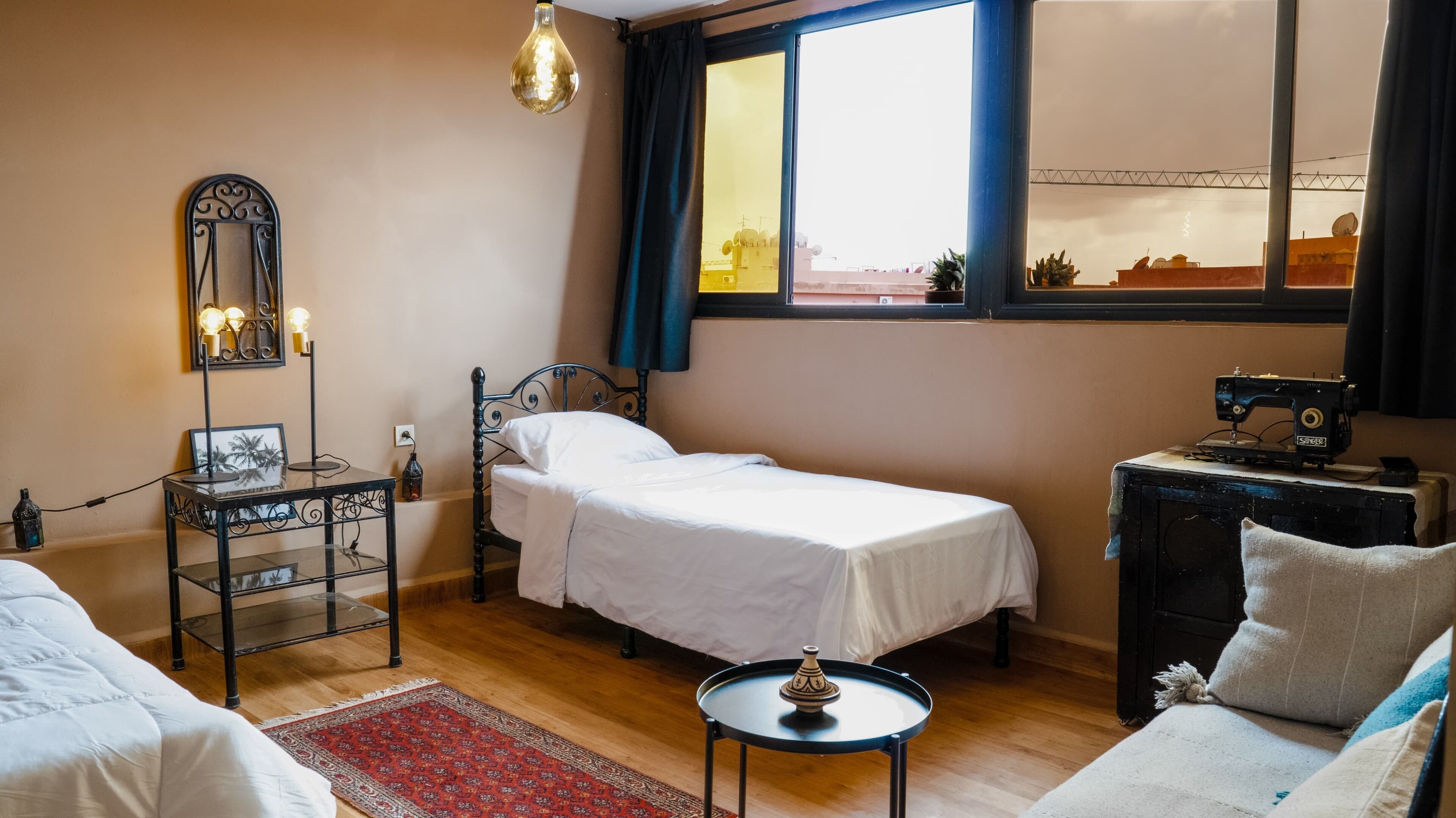A small, cozy room with a single bed, a desk with a lamp, a sewing machine, and a view of the outside through a window.