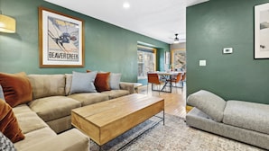 Una sala de estar con un sofá seccional color beige, una mesa de centro de madera y un sofá gris. Hay un cartel enmarcado en la pared y un comedor al fondo.