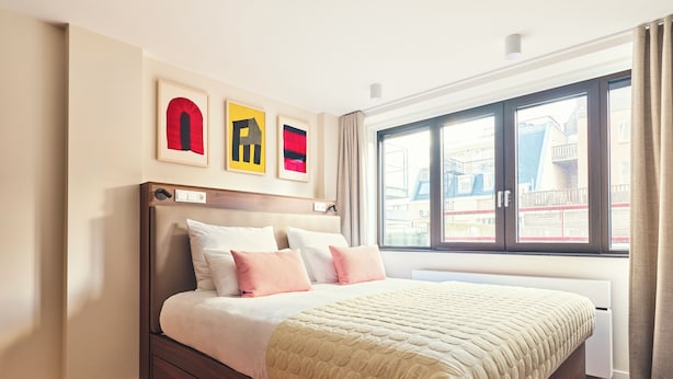 Une chambre moderne avec un grand lit, une tête de lit en bois et une vue sur les immeubles par une grande fenêtre.