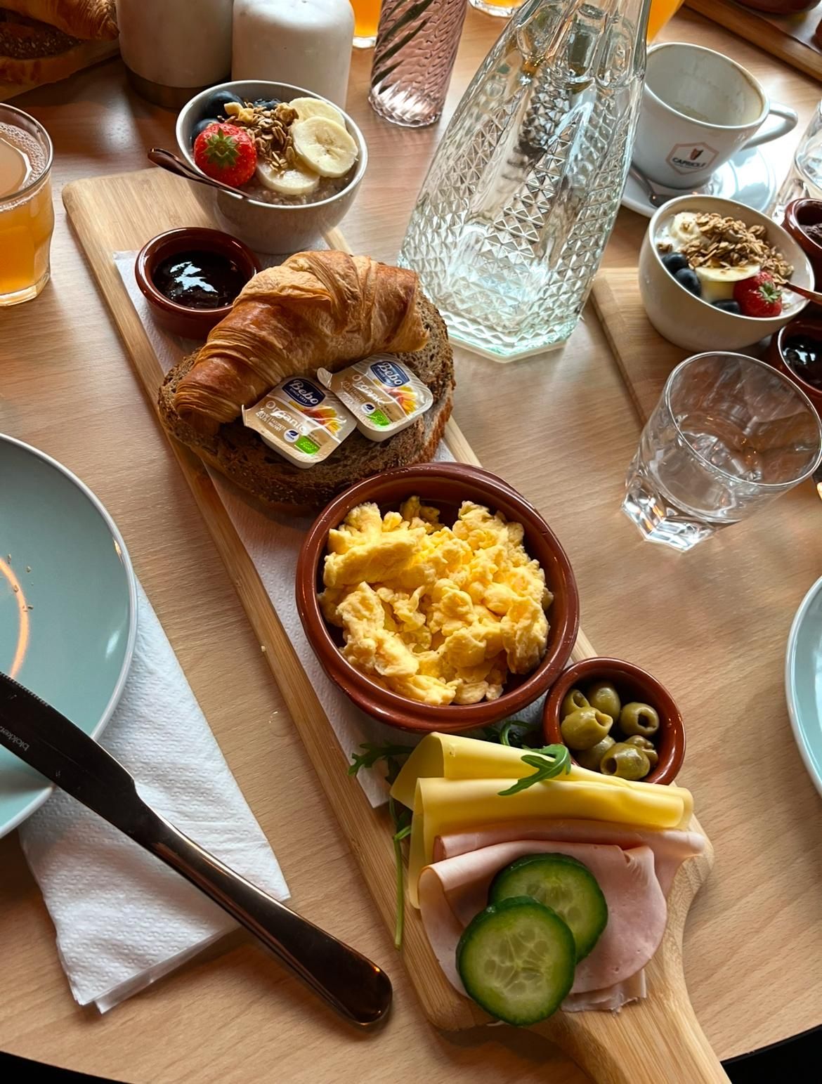 Una mesa de desayuno con cruasanes, huevos revueltos, jamón, queso, pepino, aceitunas y un vaso de zumo de naranja.