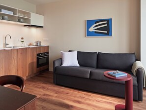 A modern living space with a kitchen, a grey sofa, a small round table, and a blue artwork on the wall. - Numa The Hague Scheveningen (The Hague)